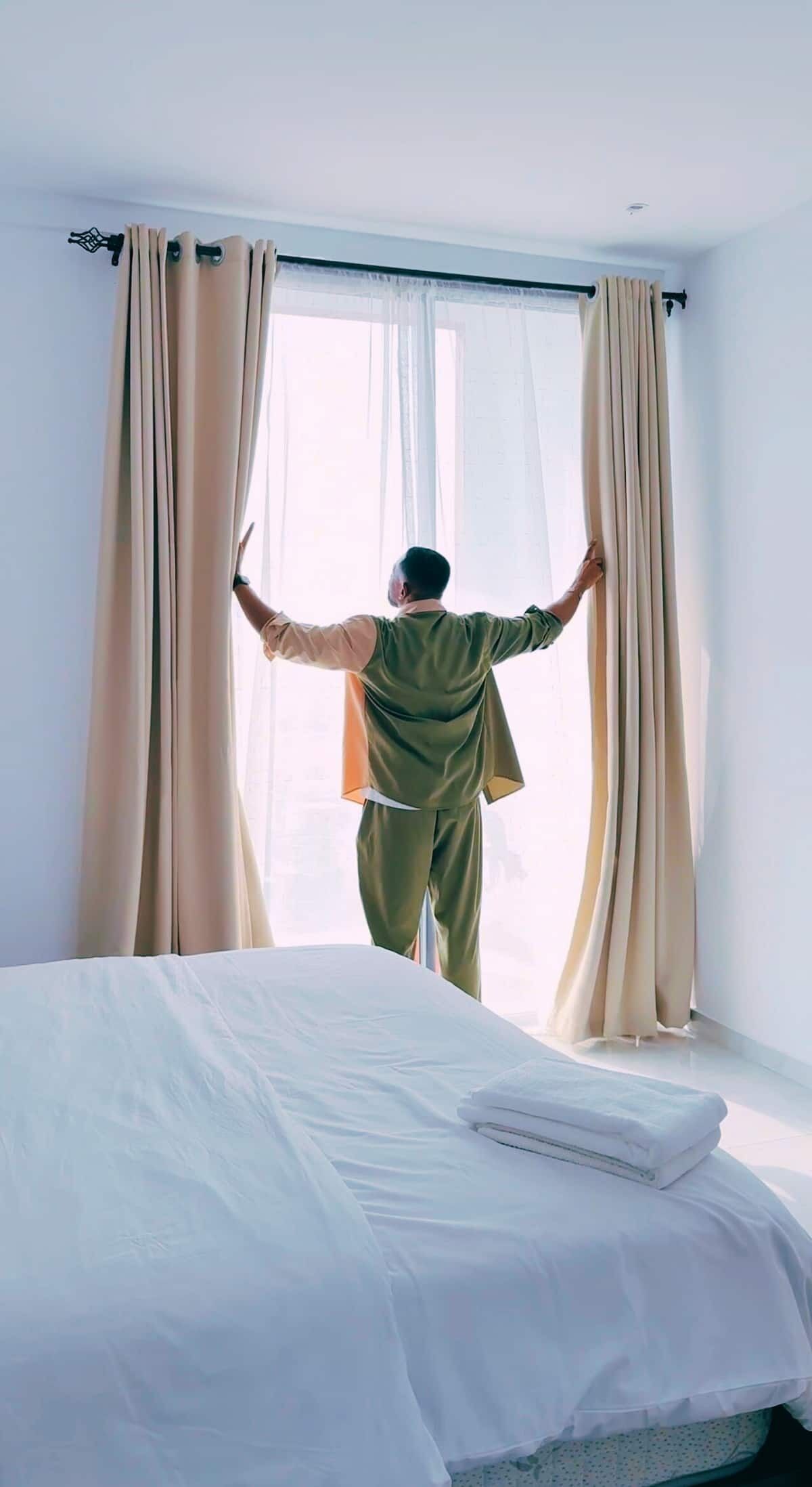 Eine Person steht in einem Zimmer mit einem Bett, Vorhängen und einem Fenster.