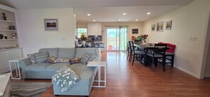 A living room with a grey sofa, a dining table with chairs, and a kitchen in the background. - Quite peaceful 3 Bedroom house in Harrington with relaxing outdoor setting. (Harrington)