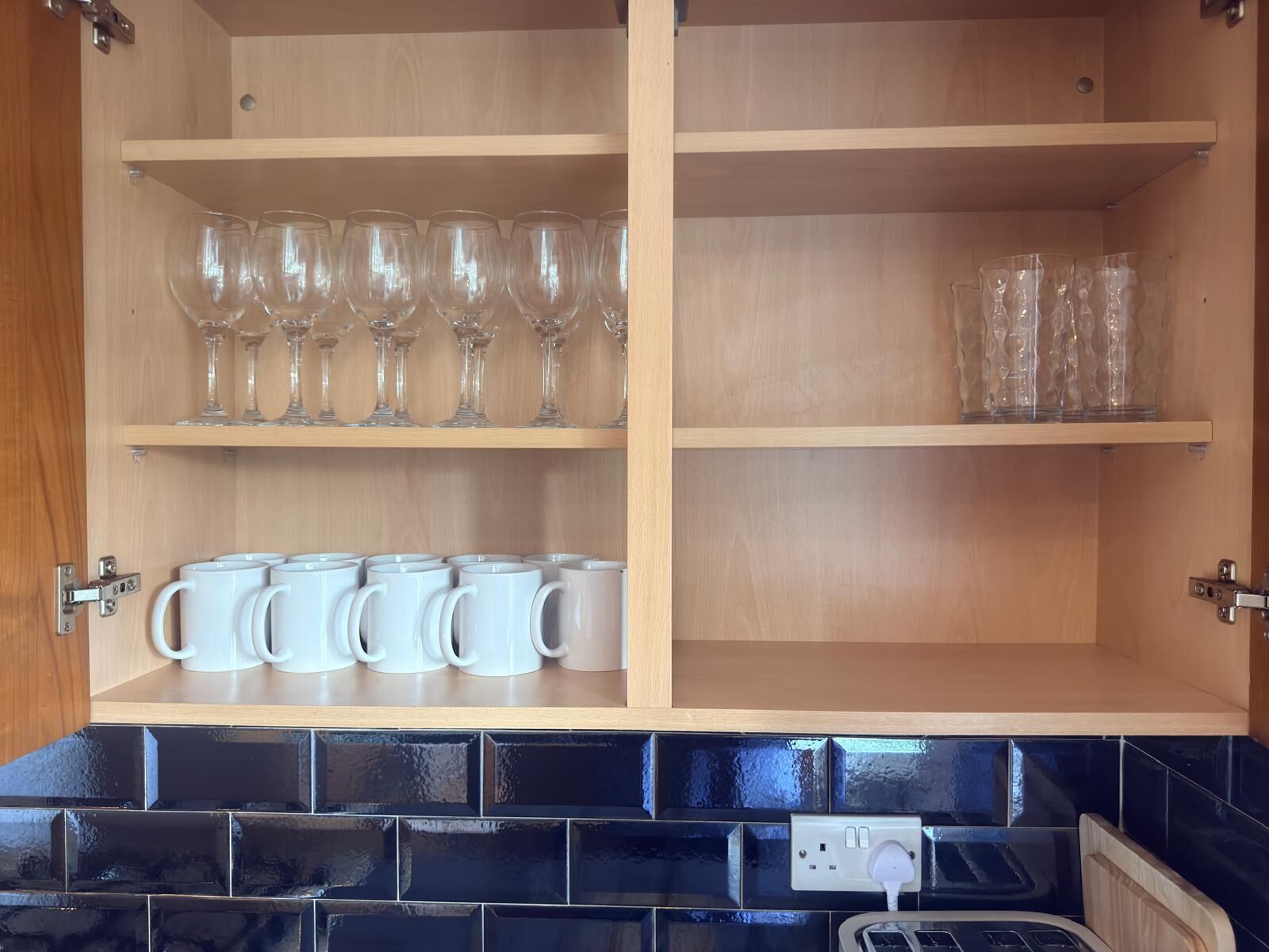 A kitchen cabinet with shelves holding wine glasses and white mugs.