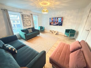 A modern living room with a sofa, a pink ottoman, a green armchair, and a television on the wall.