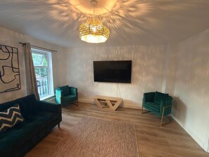 A modern living room with a large flat-screen TV, two green armchairs, a dark sofa, and a patterned rug.