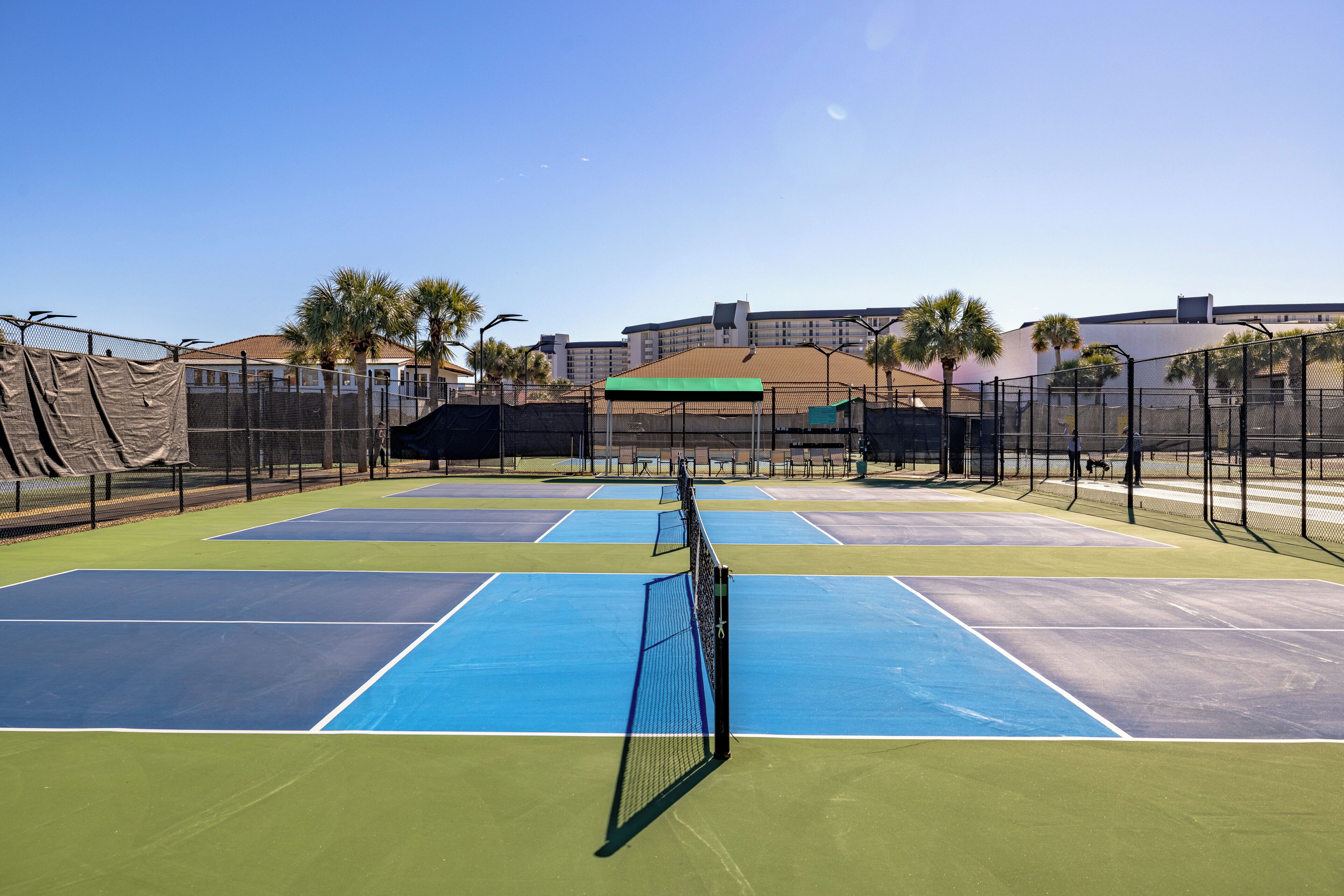 A tennis court with multiple courts, surrounded by a fence, and buildings in the background.