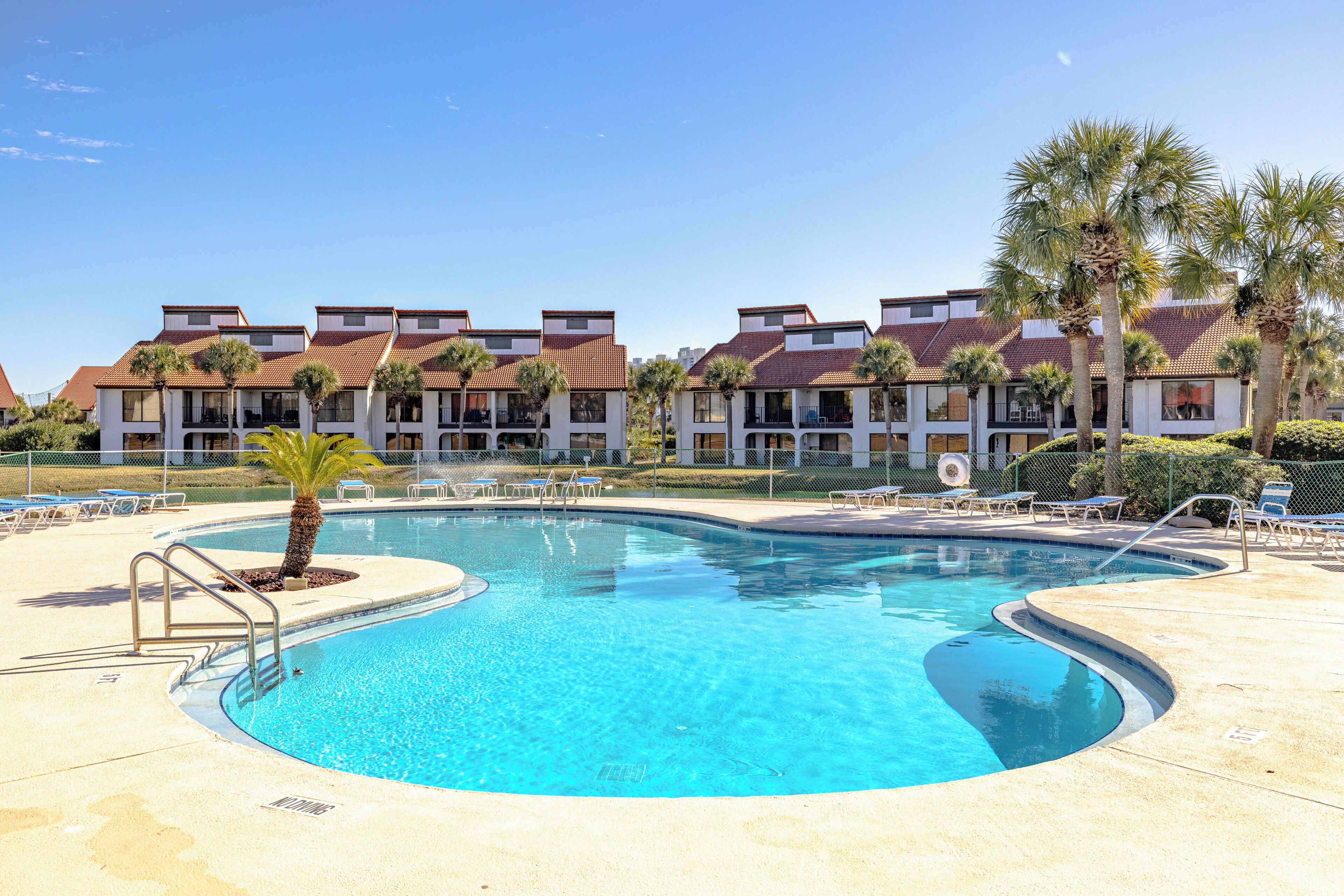 A swimming pool with a palm tree, lounge chairs, and a building with a tiled roof in the background.