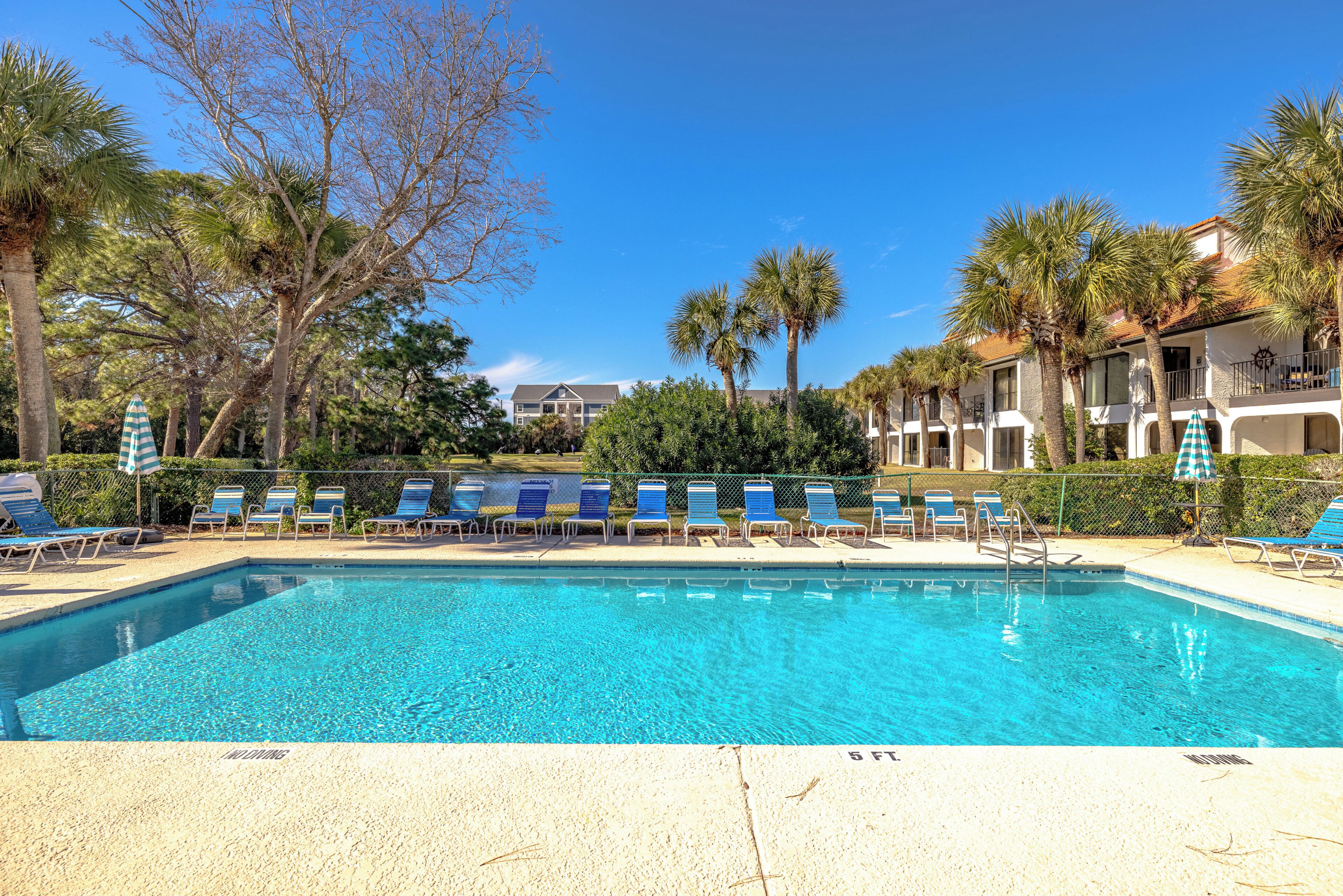 A clear blue pool with lounge chairs and umbrellas, surrounded by palm trees and a building with balconies.