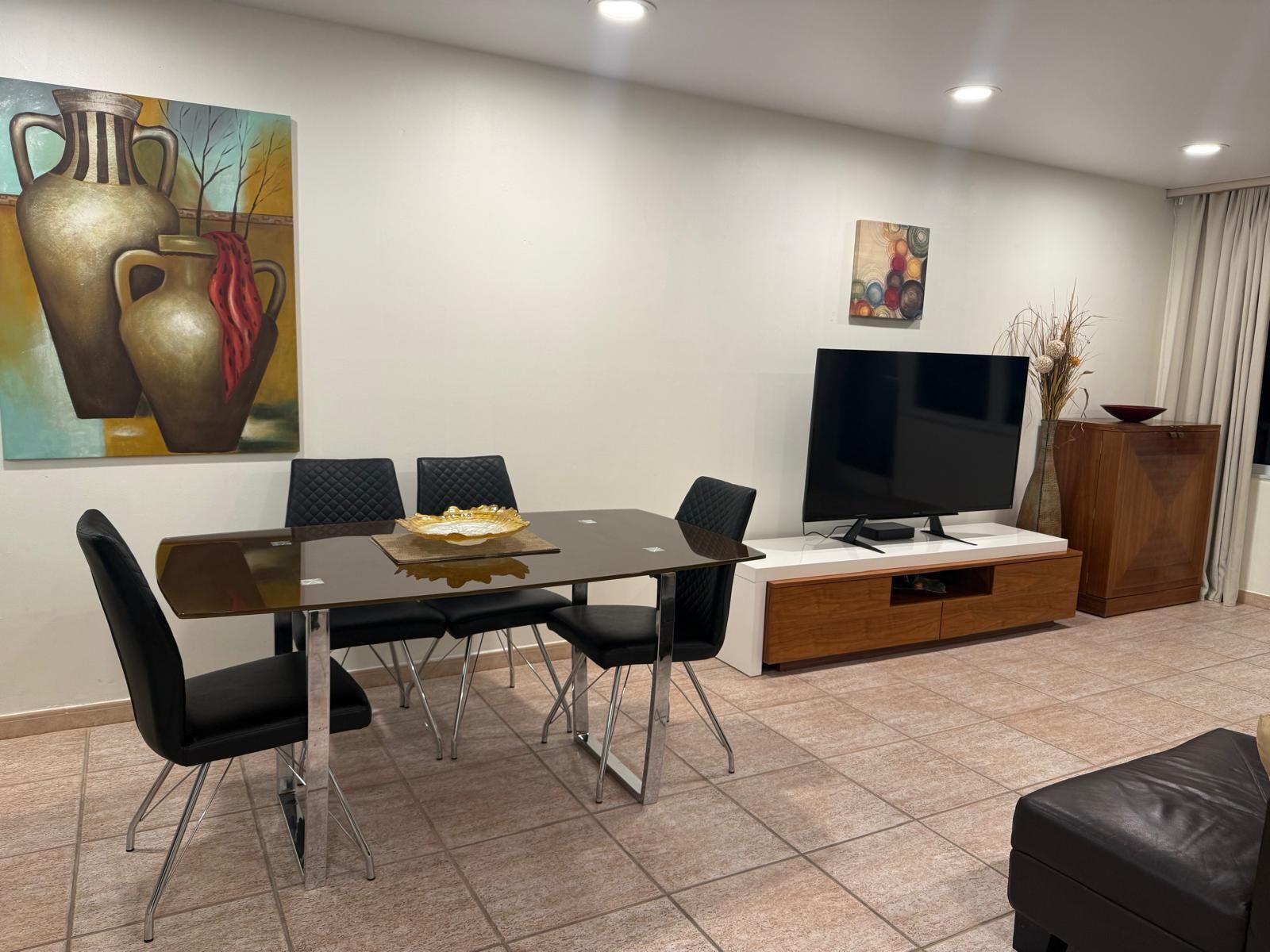 A modern living room with a dining table, chairs, a flat-screen TV, a cabinet, and artwork on the walls.