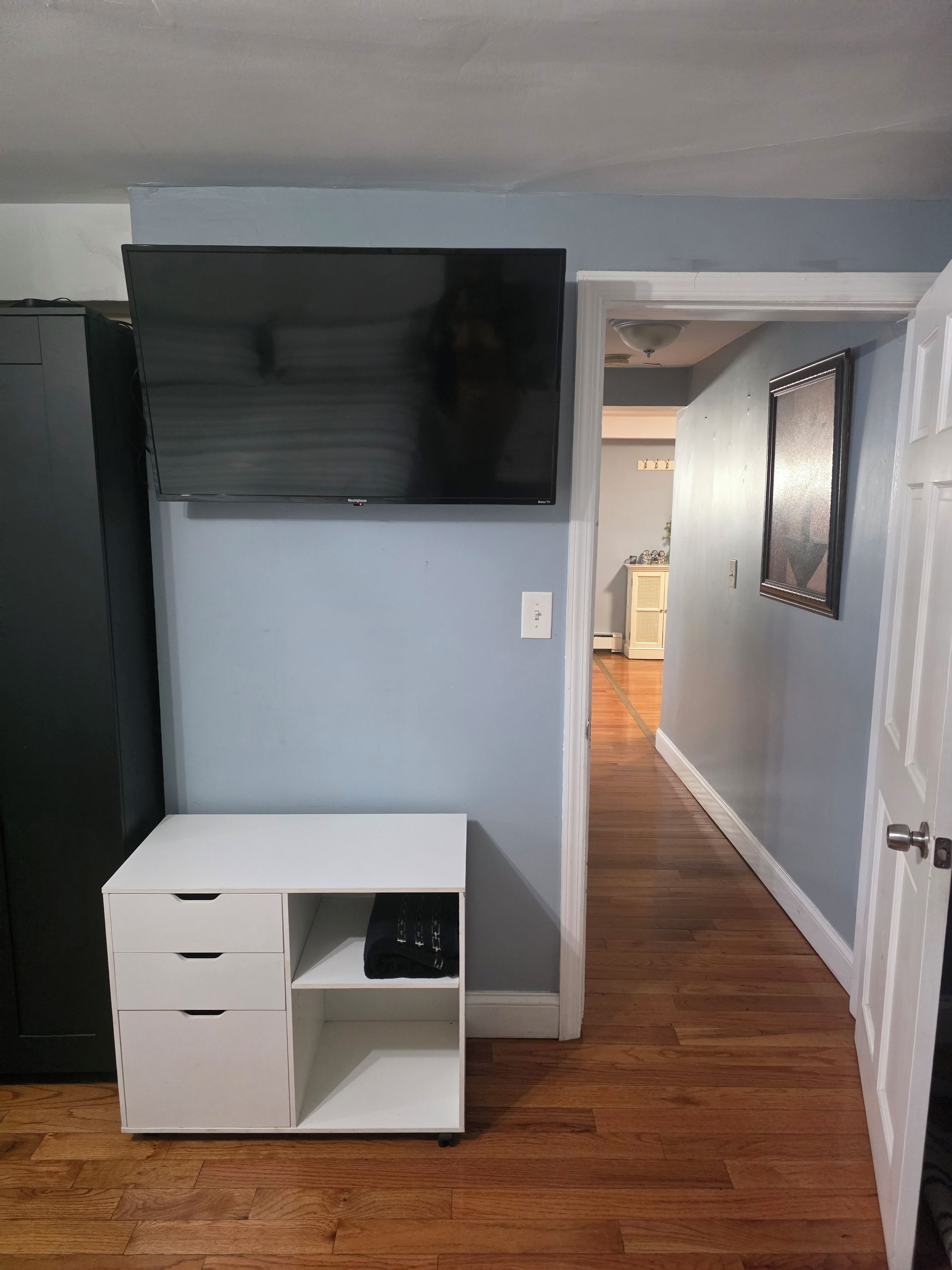 Une pièce avec un bureau blanc, un téléviseur à écran plat fixé au mur et une porte donnant sur une autre pièce.
