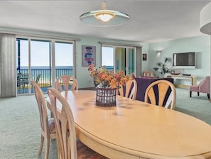 A dining area with a round table, chairs, a vase of flowers, a television, and a balcony with a view. - Spacious 3-bedroom condo with amazing Beach front views of the Gulf! (Sarasota)