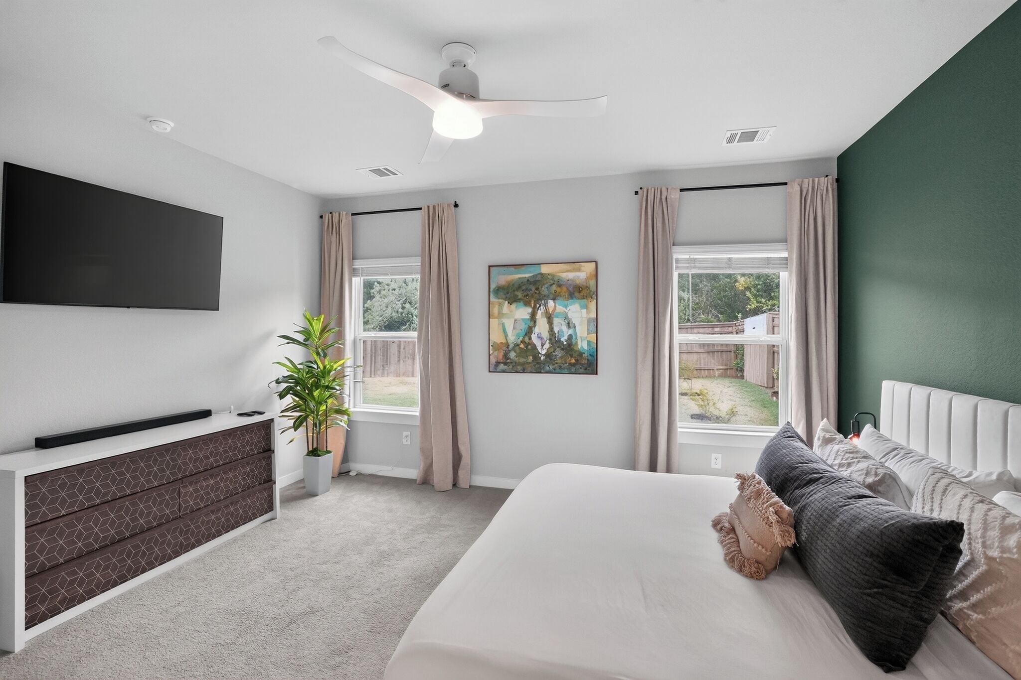 Un dormitorio con cama, televisor montado en la pared, ventilador de techo, ventana con cortinas y un cuadro.