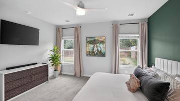 Un dormitorio con cama, televisor montado en la pared, ventilador de techo, ventana con cortinas y un cuadro.