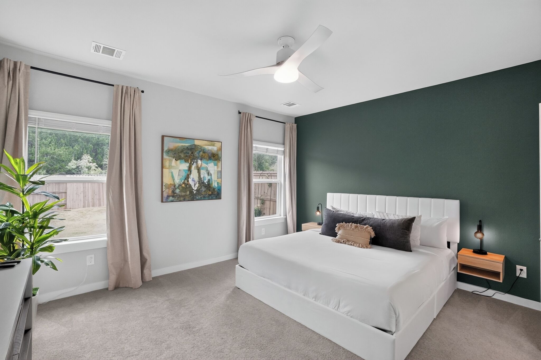 Un dormitorio con cama, mesita de noche, lámpara, ventana con cortinas, ventilador de techo y una obra de arte en la pared.