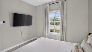 A bedroom with a bed, a flat-screen TV mounted on the wall, a window with curtains, and a ceiling fan.