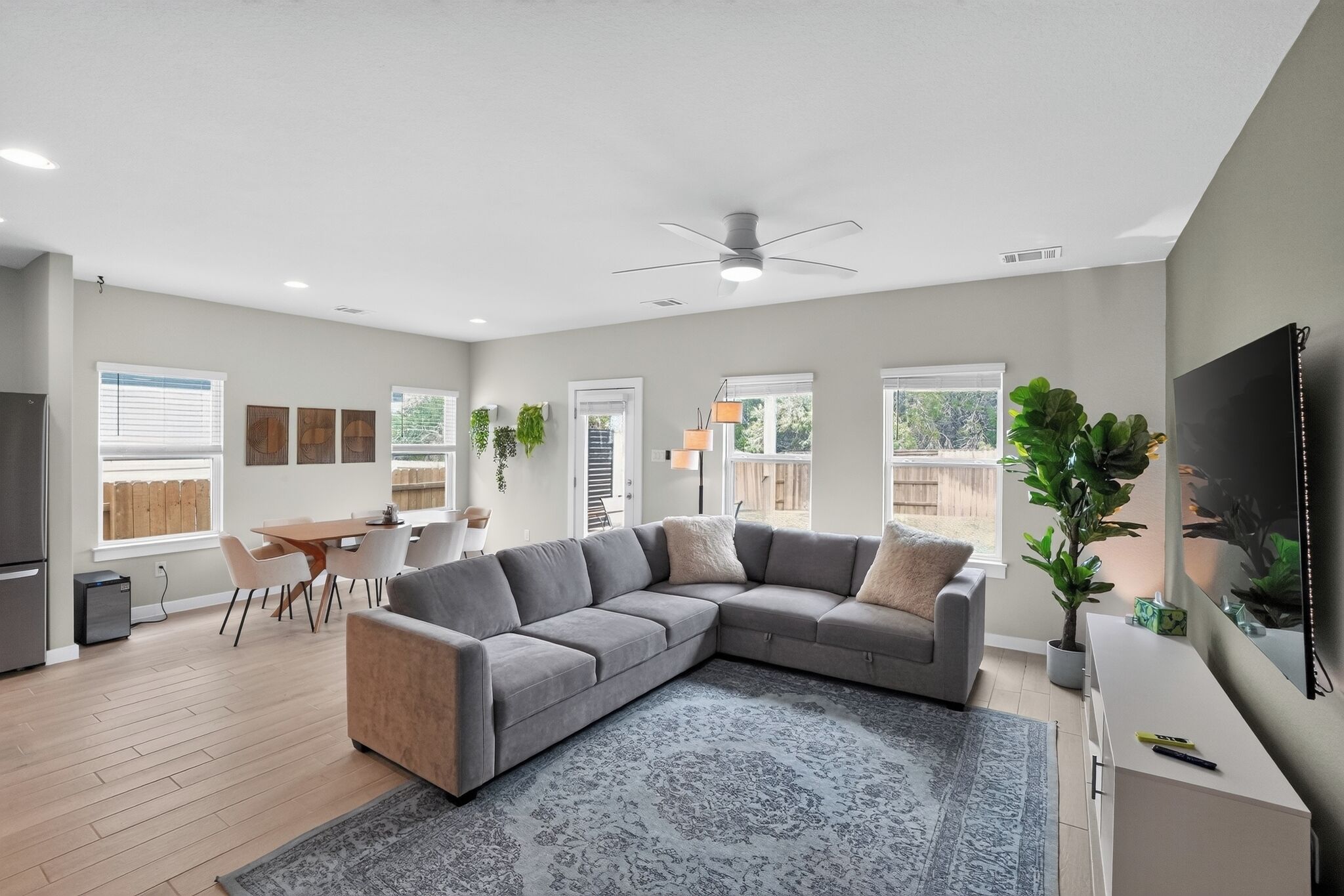 A modern living room with a grey sectional sofa, a dining area with a table and chairs, a kitchen area with a fridge, and a large window providing natural light.