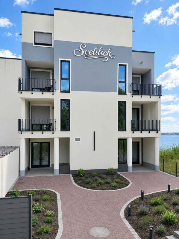 A modern multi-story building with a sign reading 'Seeblick.' It features balconies, large windows, and a landscaped entrance area.