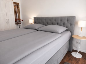 A neatly made bed with grey linens and a tufted headboard, accompanied by two bedside lamps on a round wooden table.