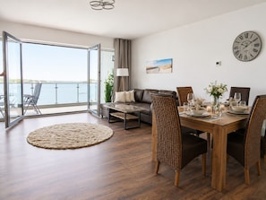 A modern living room with a dining area, a sofa, a coffee table, and a view of the sea through a glass door.