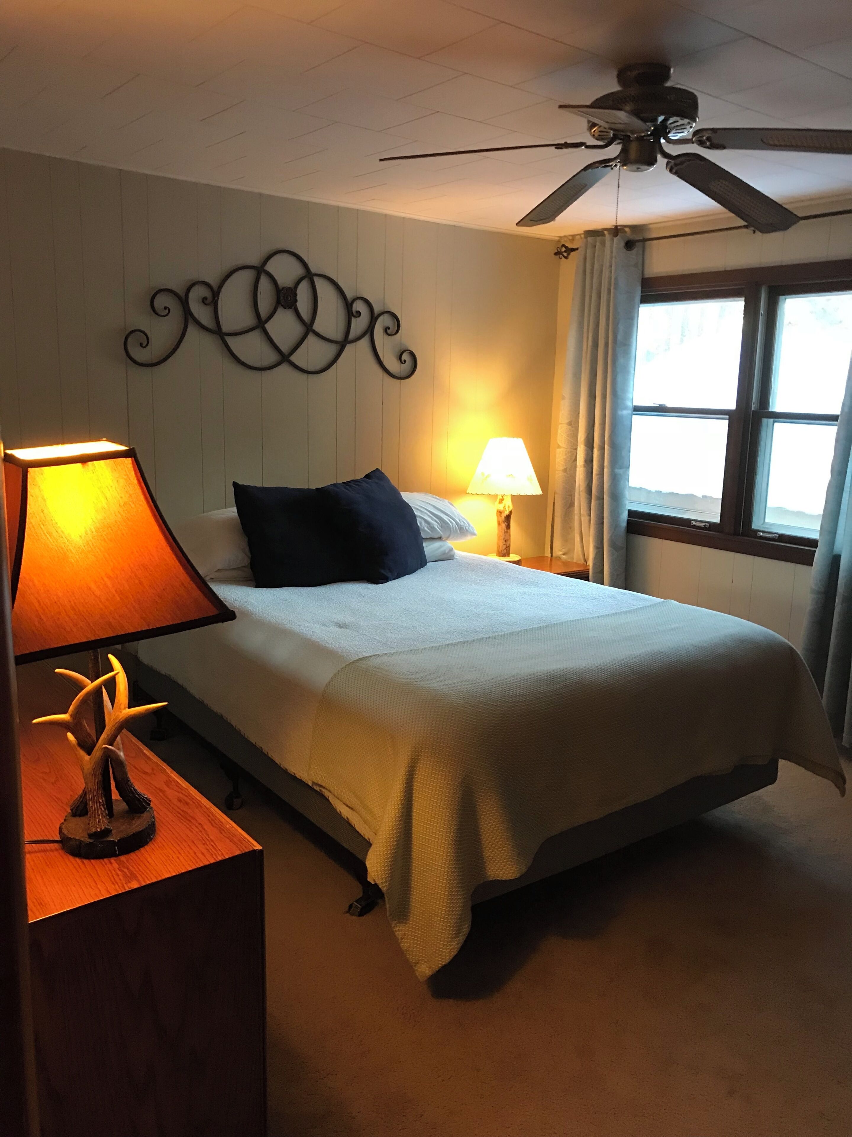 A bedroom with a bed, a nightstand with a lamp, a ceiling fan, and a window with curtains.