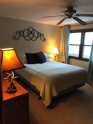 Une chambre à coucher avec un lit, une table de chevet avec une lampe, un ventilateur de plafond et une fenêtre avec des rideaux.