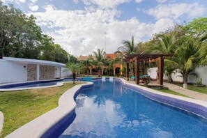 A swimming pool with a curved edge, surrounded by grass and palm trees, with a building in the background. - Villa Coral (Tulum)