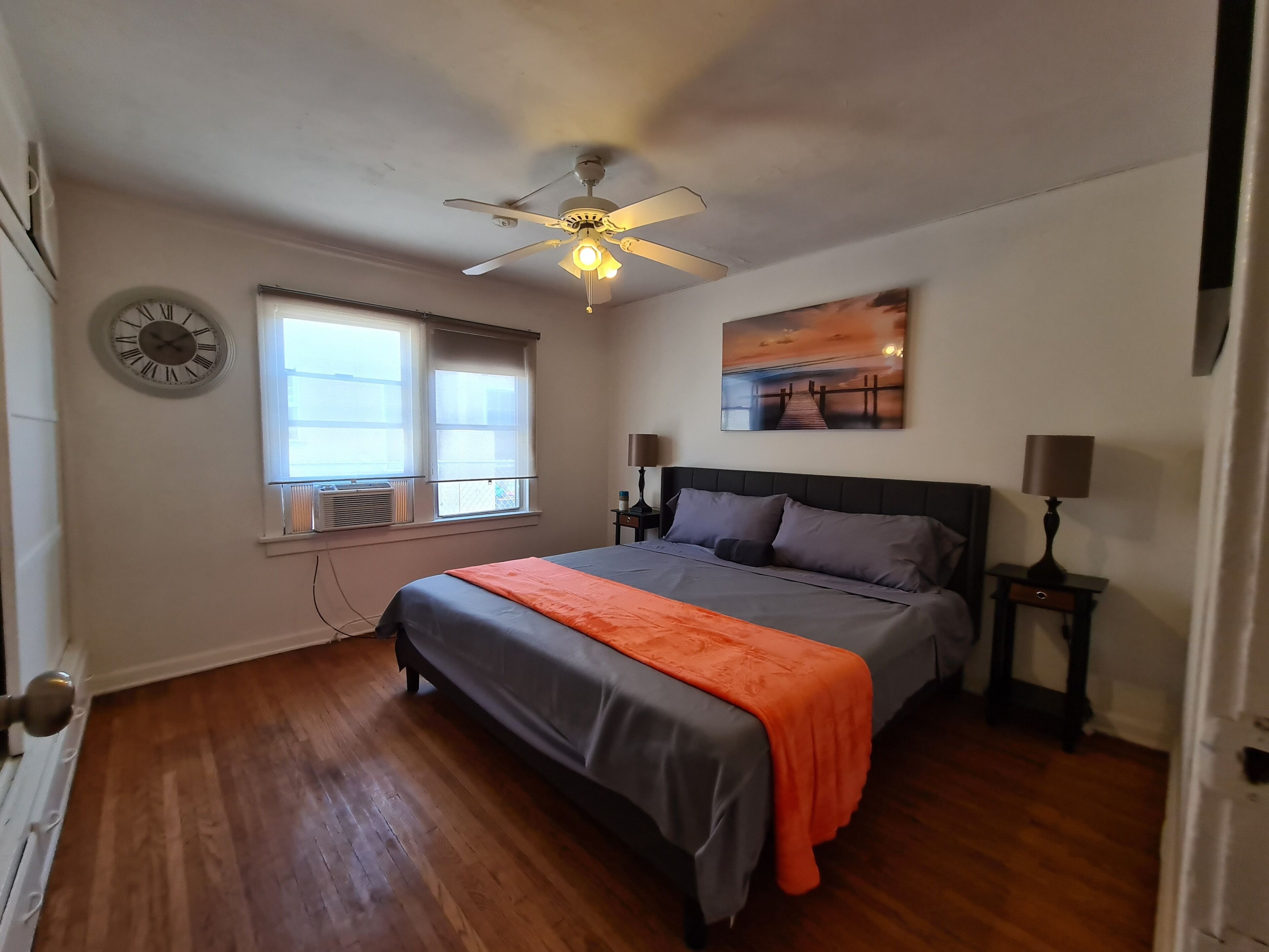 A bedroom with a bed, a ceiling fan, a window with blinds, a wall clock, a painting, and a nightstand with a lamp.
