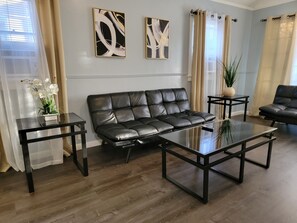 Een moderne woonkamer met een leren bank, een glazen salontafel en twee bijzettafels. Aan de muur hangen abstracte schilderijen en op een bijzettafel staat een pot met een plant.