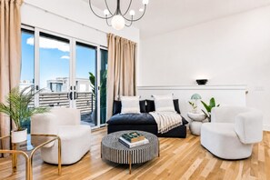 Un dormitorio moderno con un ventanal grande, una cama, dos sillones, un pouf redondo y una mesita de noche con una lámpara.