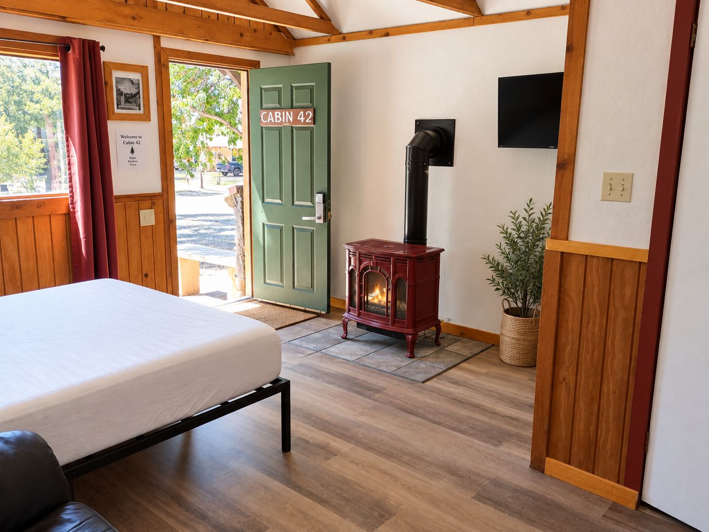 A cabin interior with a bed, a wood-burning stove, a green door labeled 'Cabin 42', and a view of the outside.