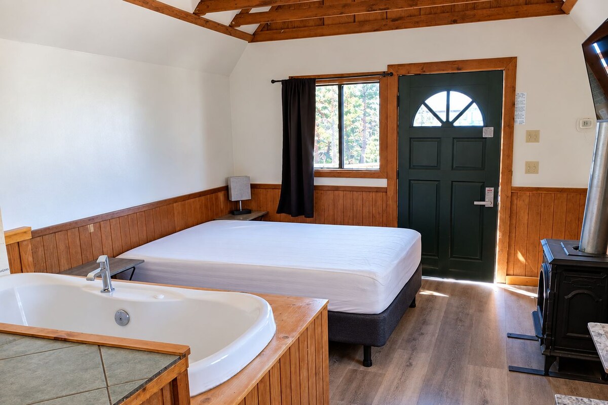 A room with a wooden floor, a large bathtub, a bed, a door, and a stove.