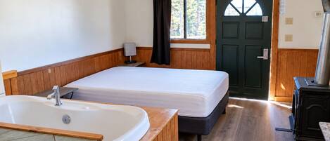A room with a wooden floor, a large bathtub, a bed, a door, and a stove.