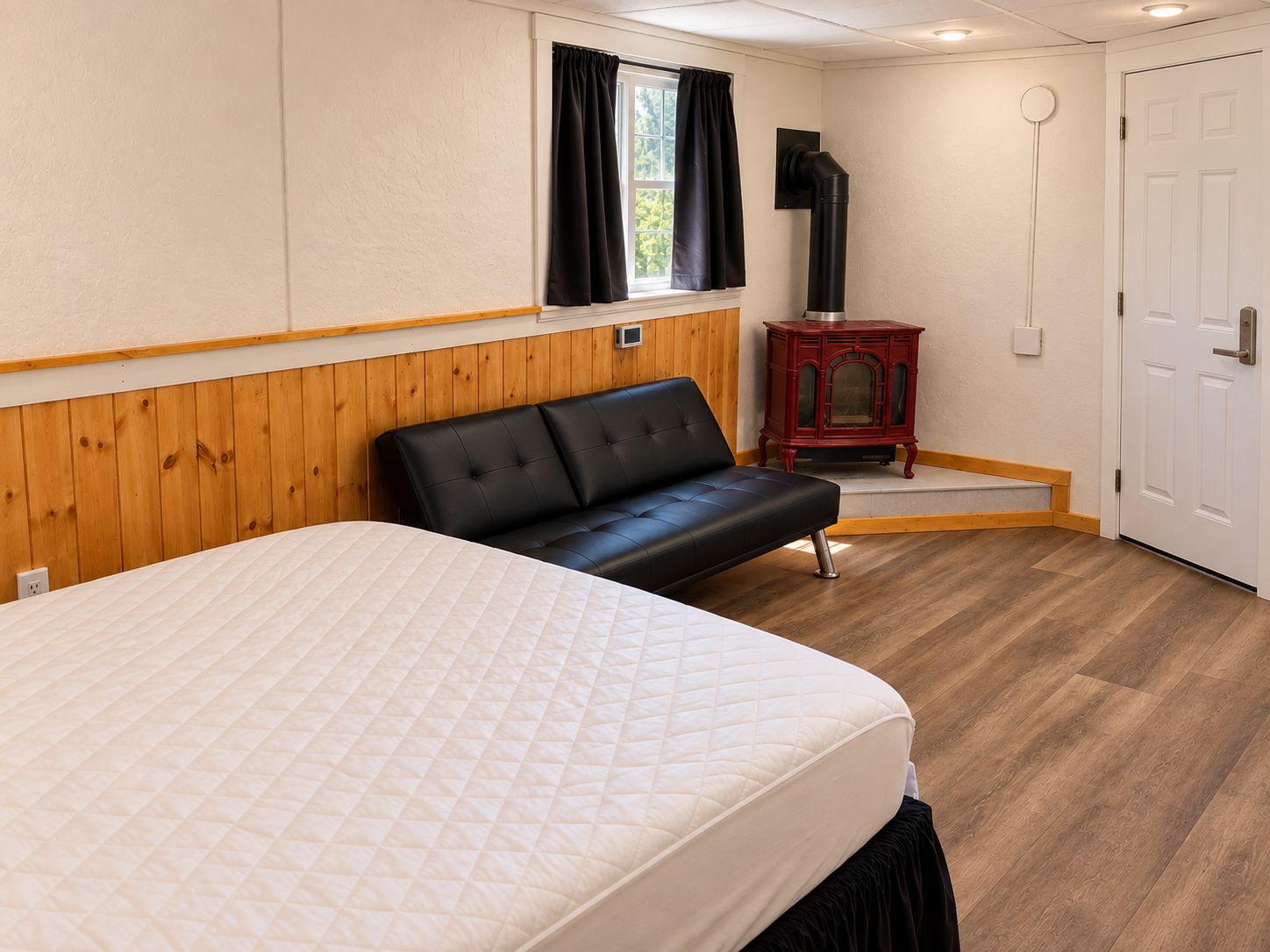Una habitación con cama, un banco, un calentador, una puerta y una ventana con cortinas.