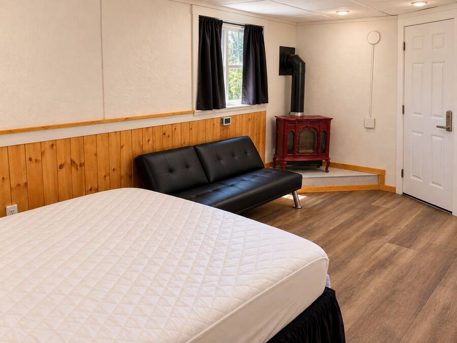 Una habitación con cama, un banco, un calentador, una puerta y una ventana con cortinas.