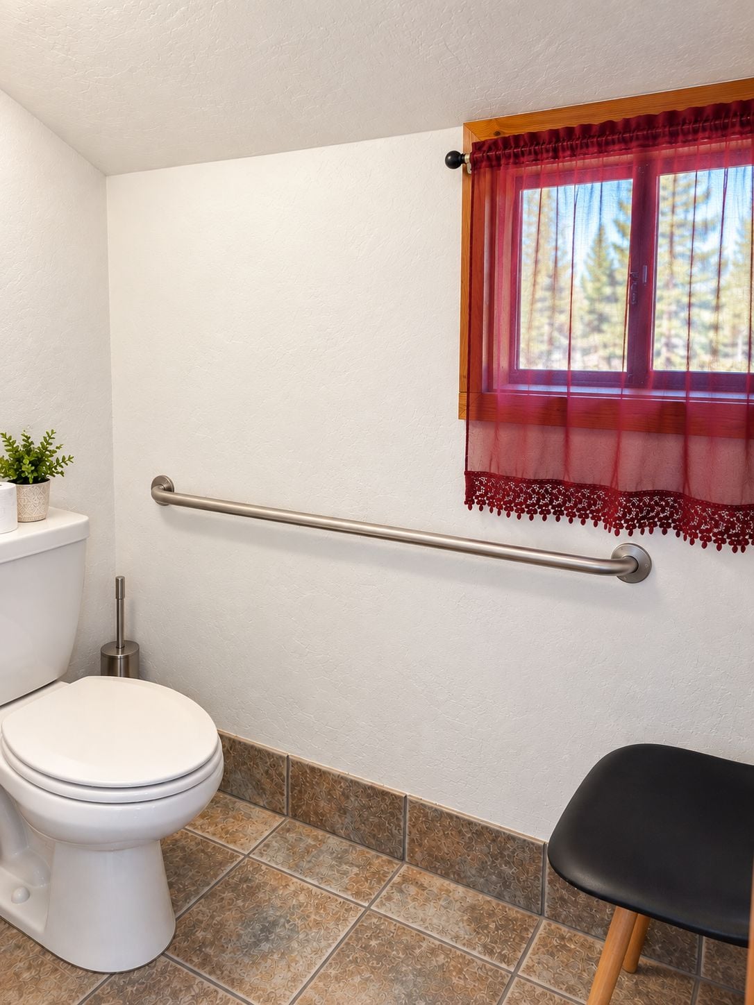 A bathroom with a toilet, a handrail, a window with a red curtain, and a potted plant.