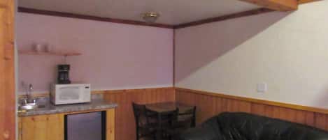 A compact kitchen with a microwave, a small refrigerator, and a sink. There is a wooden dining table with chairs and a black leather sofa.