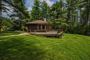 A wooden cabin with a deck, surrounded by a well-maintained lawn and dense forest.