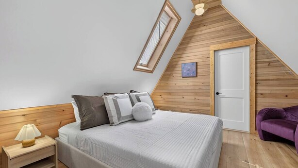 A bedroom with a sloped ceiling, a bed with pillows, a nightstand, a lamp, a door, and a purple chair.