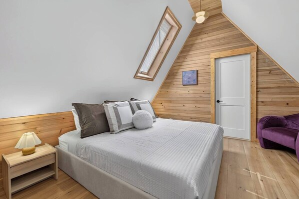 A bedroom with a sloped ceiling, a bed with pillows, a nightstand, a lamp, a door, and a purple chair.