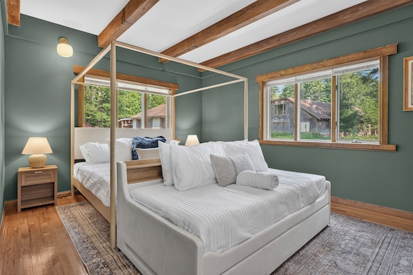 Een hemelbed met een wit hoofdeinde en voeteinde, een nachtkastje met een lamp, en een groot raam met uitzicht op een huis en bomen.