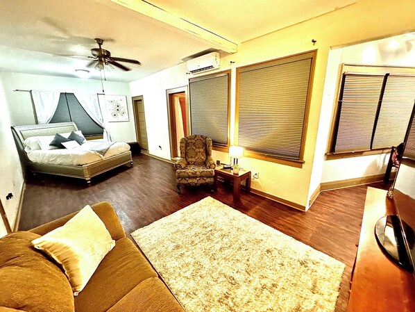 A living room with a sofa, a chair, a small table with a lamp, and a bed with a headboard.