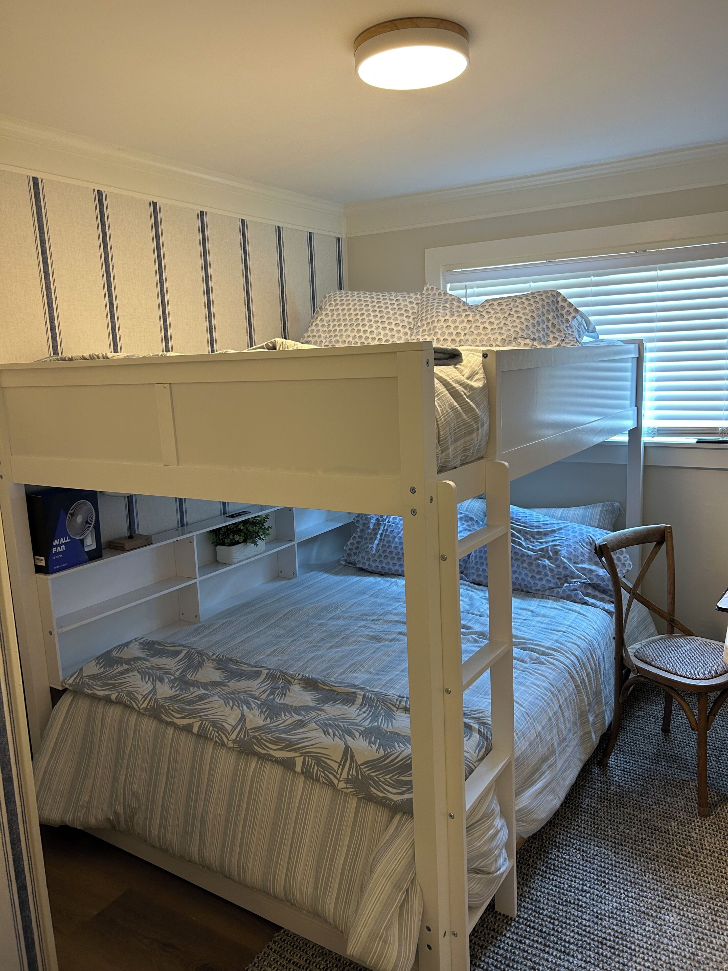 Une chambre avec un lit superposé, un ventilateur de plafond, une chaise et une fenêtre avec des stores.