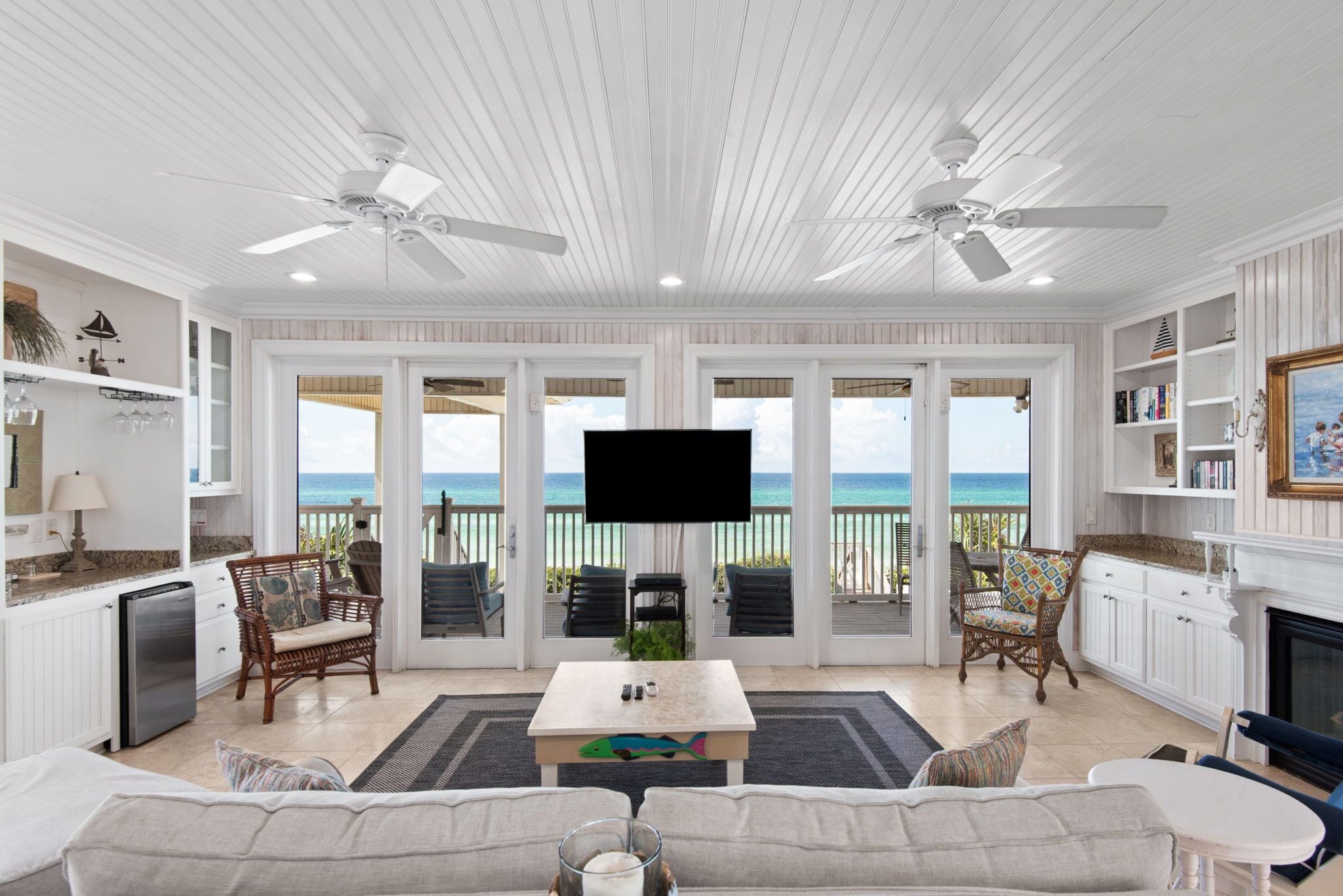 A spacious living room with a large flat-screen TV, a white sofa, and a coffee table with a fish design. There are two ceiling fans, a fireplace, and a view of the ocean through large glass doors.