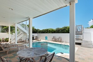 A covered outdoor swimming pool area with lounge chairs, a table, and a staircase leading to a deck.
