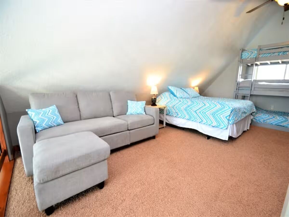 A room with a grey sofa, a bed with blue and white bedding, a small table with a lamp, and a bunk bed.