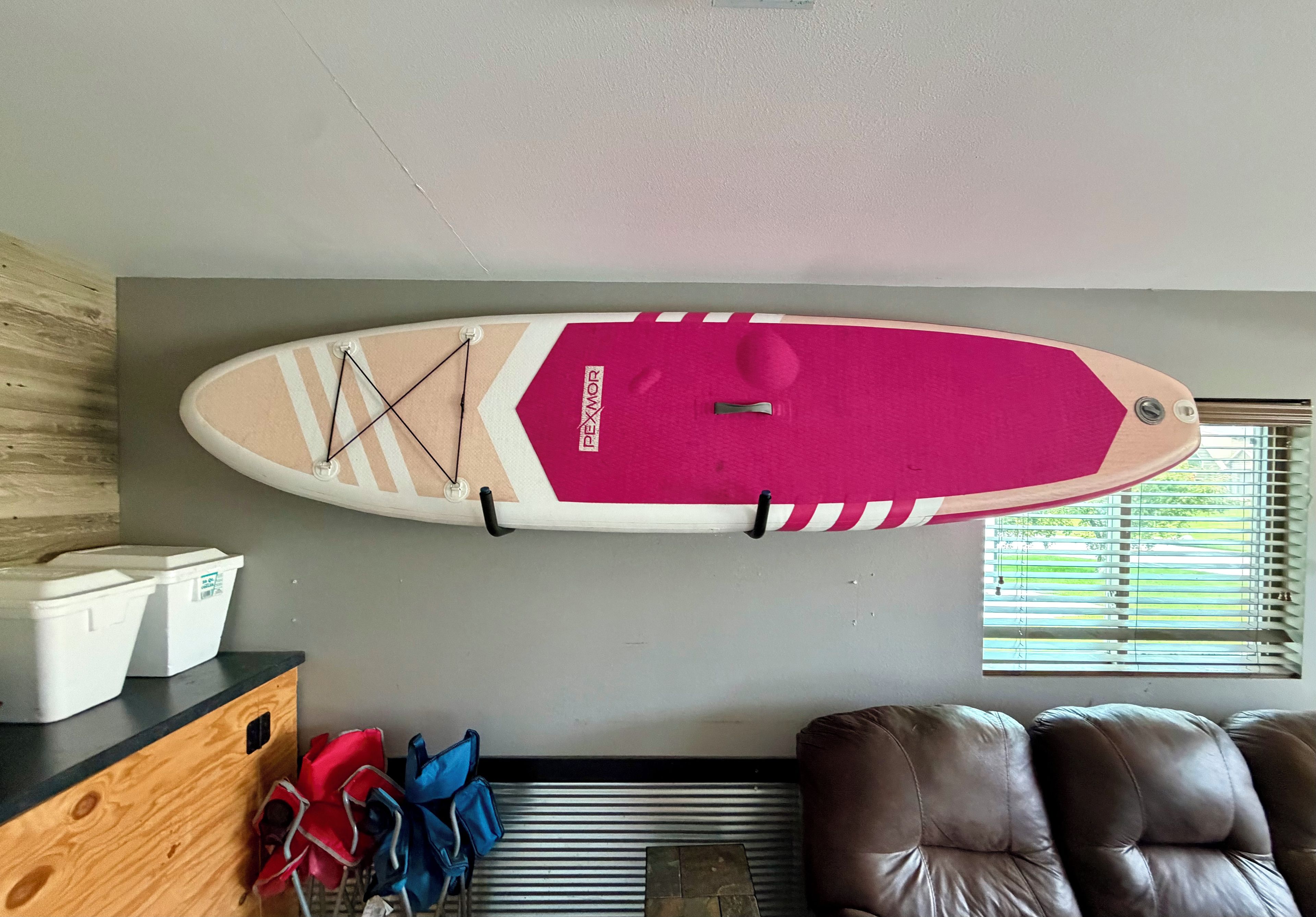 A ceiling-mounted surfboard with a pink and white design, a wooden cabinet with storage bins, and a brown leather couch.