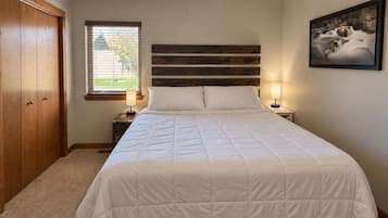Une chambre à coucher avec un lit, une tête de lit en bois, deux lampes de chevet, un ventilateur de plafond, une fenêtre avec des stores et un tableau encadré au mur.