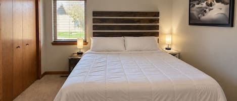 Ein Schlafzimmer mit einem Bett, einem Holz-Kopfteil, zwei Nachttischlampen, einem Deckenventilator, einem Fenster mit Jalousien und einem gerahmten Bild an der Wand.