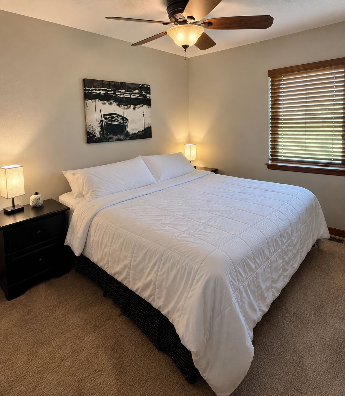 A neatly made bed with white bedding, a ceiling fan, a wall-mounted painting, and a window with blinds.