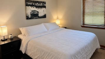 A neatly made bed with white bedding, a ceiling fan, a wall-mounted painting, and a window with blinds.