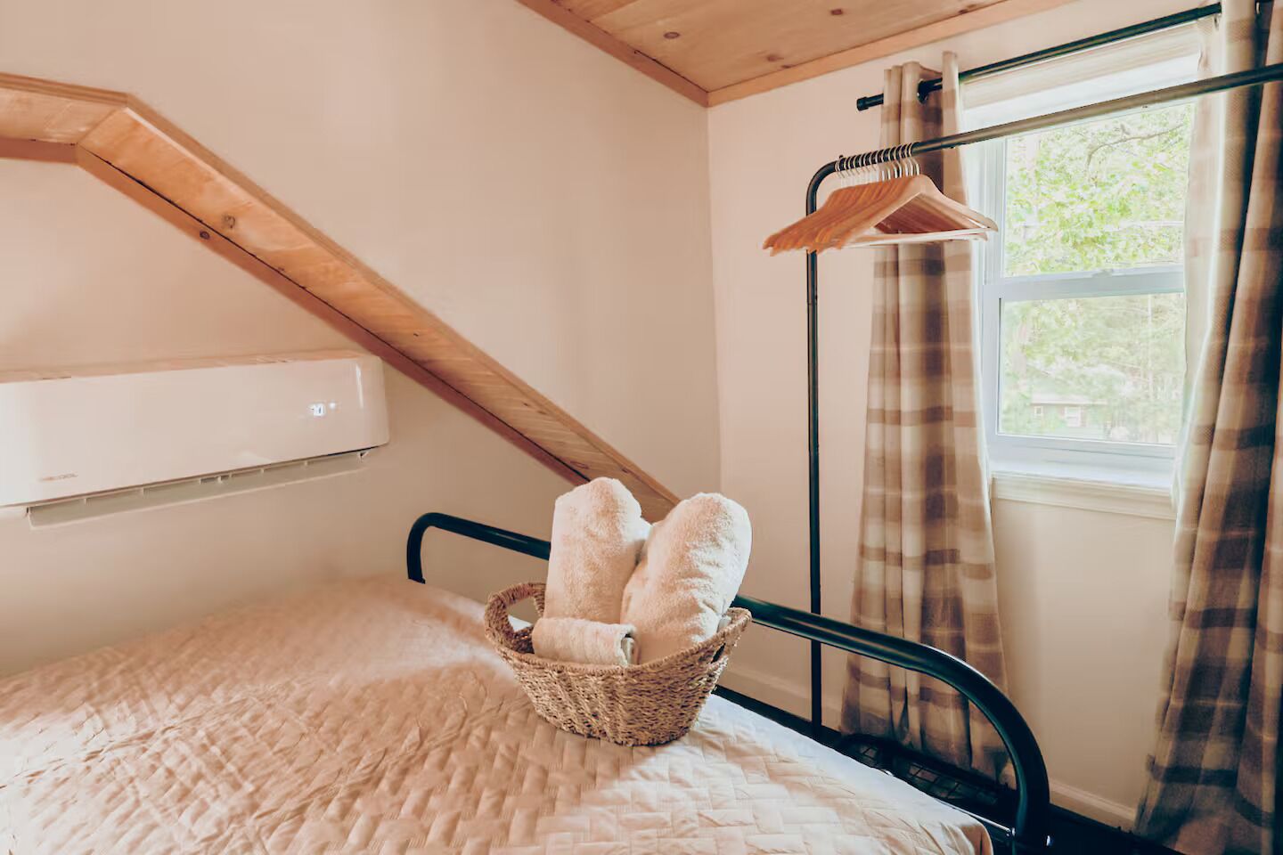 Una camera da letto con soffitto a falde, un letto con un cesto intrecciato pieno di asciugamani arrotolati, una finestra con tende e un condizionatore d'aria a parete.