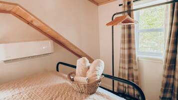 Una camera da letto con soffitto a falde, un letto con un cesto intrecciato pieno di asciugamani arrotolati, una finestra con tende e un condizionatore d'aria a parete.