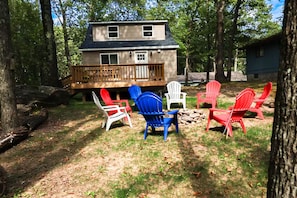 A two-story cabin with a deck, surrounded by trees. There are several Adirondack chairs in various colors.