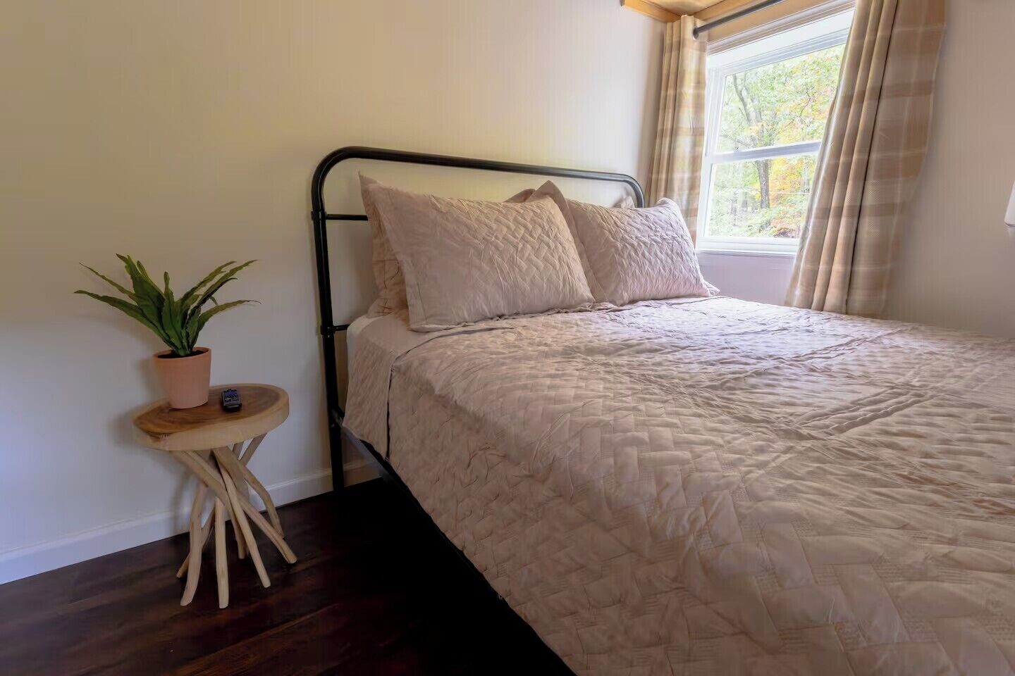 A bed with a quilted cover, a bedside table with a potted plant, and a window with curtains.